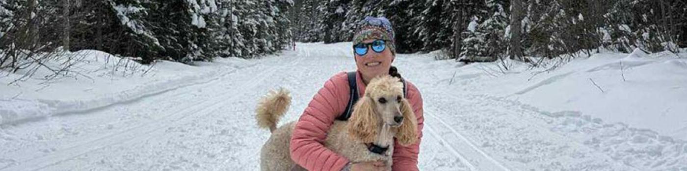 Haylee holding her dog on one knee in the snow, surrounded by trees