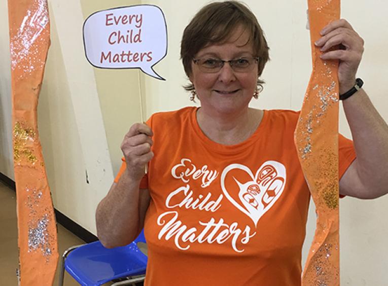 A middle-aged woman wearing an Orange Shirt Day shirt that says "every child matters" holds an orange frame and a sign. The sign also says, "every child matters".