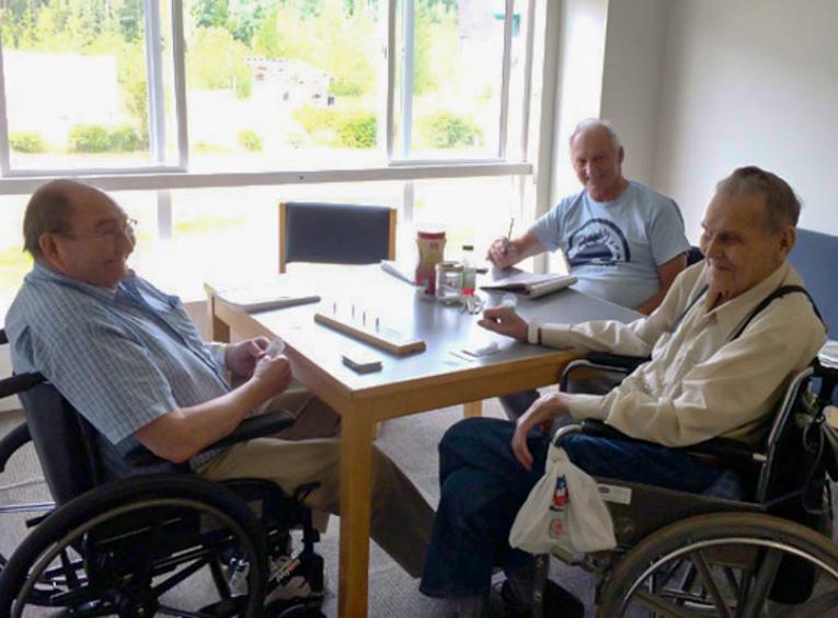 Three elderly men sitting around a table, two in wheelchairs