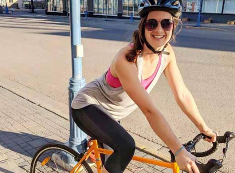A woman wearing a bike helmet, perched on an orange bicycle, on a sidewalk downtown.