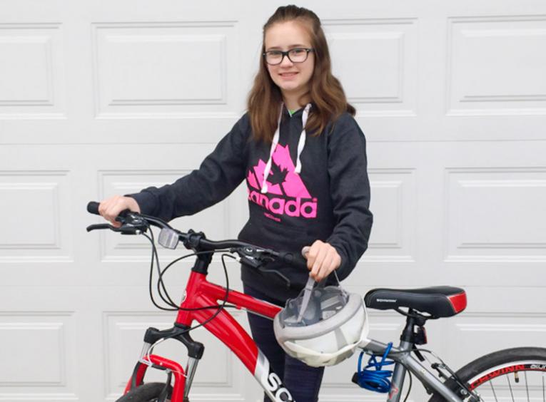 A young girl, wearing a black and pink hoodie, poses with her red bike and helmet.