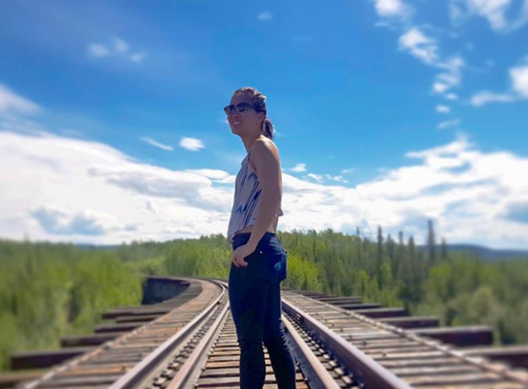 Melinda stands on a train track that disappears in the distant forest. A sunny sky beats down on her.