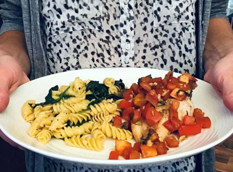 A person holds a white plate of food. On the left of the plate is pasta noodles with spinach, on the right is a chicken breast covered in chunks of tomato.