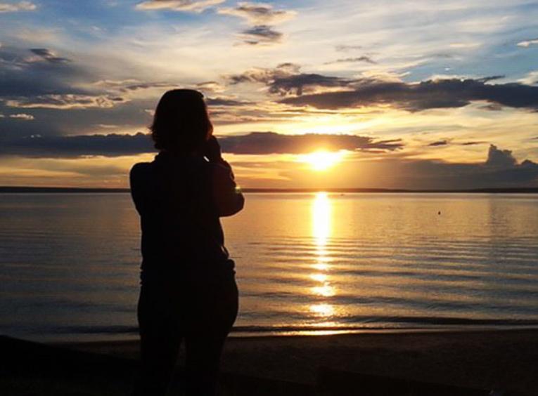 The sun sets over water in the distance. The sky is blue and gold punctuated by clouds. In the foreground, a silhouette watches the beautiful scene.
