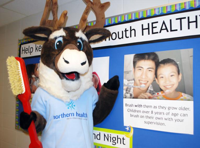 Spirit the Cariboo holding a toothbrush with a poster that shows how adults should brush with children.