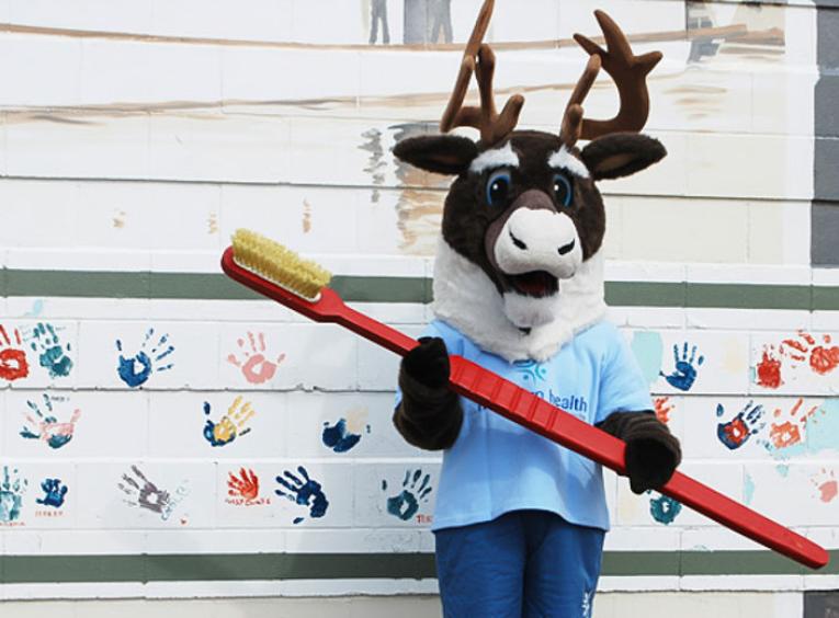 Spirit the Cariboo holding a large toothbrush, standing against a mural of handprints.