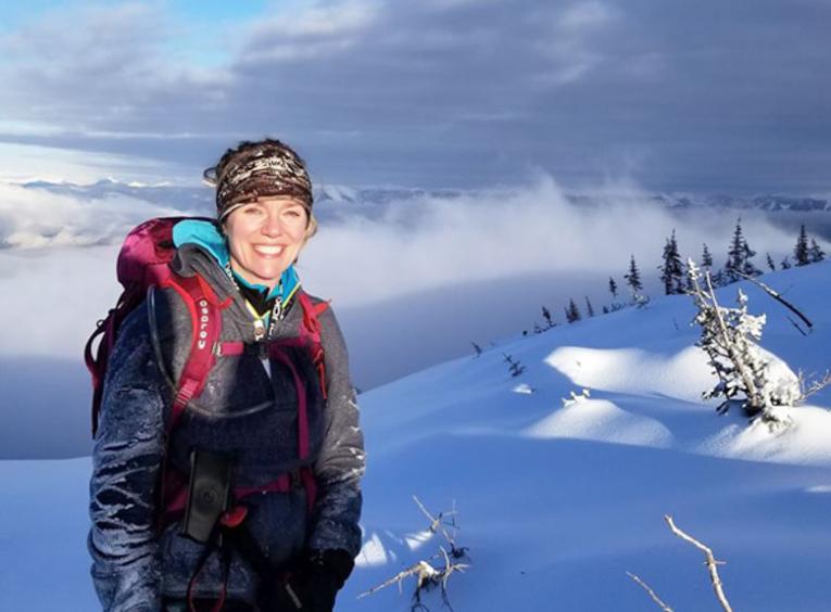 Tanya Carter is pictured on top of a mountain in the snow. In the distance is a snowy mountain range.