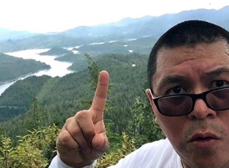 Man with index finger in the air with river and mountains in the background.