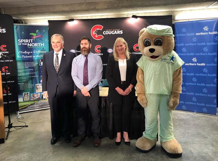 A man in a suit, a man in a tie, a woman in a suit, and a giant teddy bear mascot lined up.