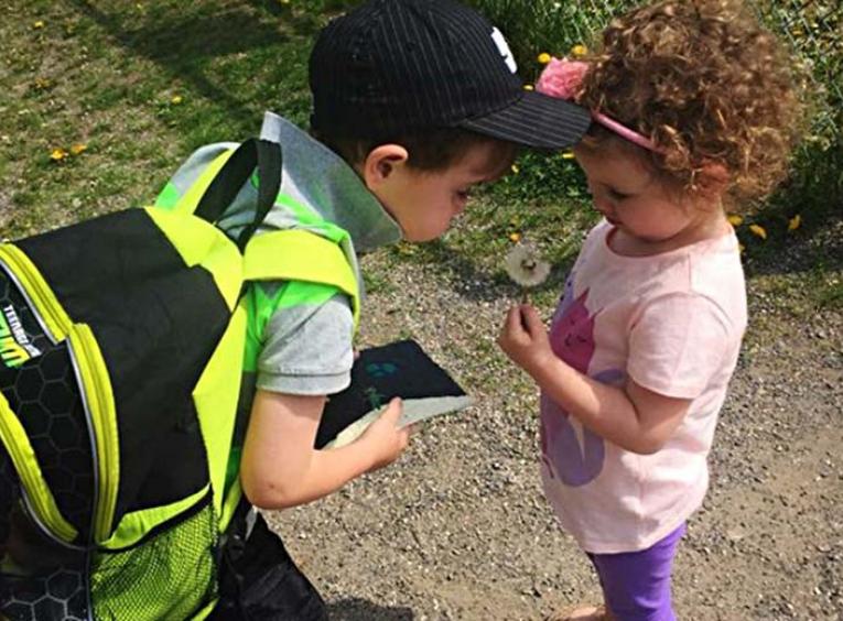 Young boy with backpack and ball cap on interacting with another young child.