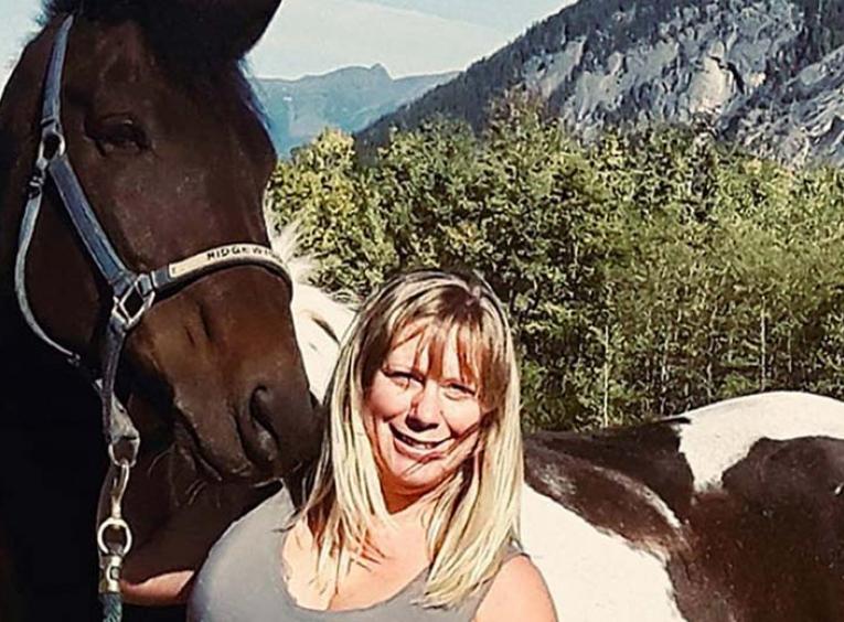 Clare smiles into the camera. Her brown and white horse is directly behind her. Mountains and forest is further in the background.