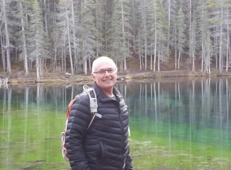 Man with white hair and glasses wearing a black jacketstanding in front of a lake in the woods.