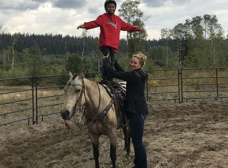 A woman poses, holding the legs of a young boy who is standing on top of a saddled horse in a field.
