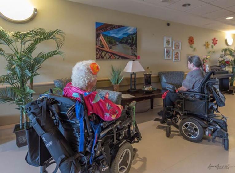 Two female Gateway Lodge residents in motorized wheelchairs in a hallway admiring art hanging on the wall.