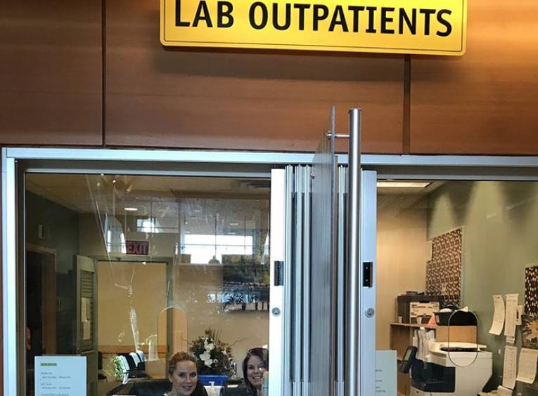 Two women behind the glass partition in a reception area under a sign that says Lab Outpatients.