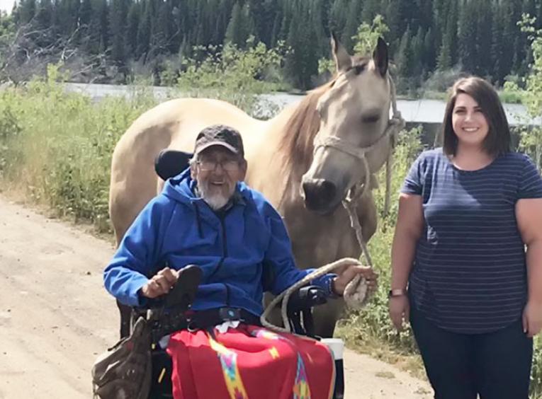 On a dirt road with a creek in the background, a man in a motorized wheelchair holds a beige horse. Woman stands to their right.