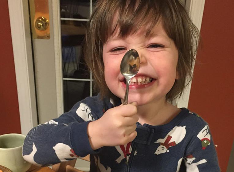 A young, smiling boy wearing polar bear pyjamas and sitting at a kitchen table holds a spoon up to his nose.