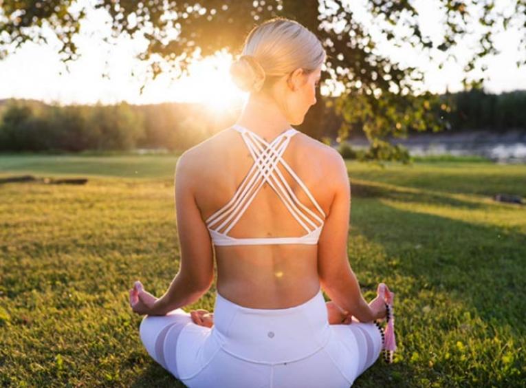 Woman practising yoga, sitting cross-legged on grass with her back to the camera. The sun sets in the horizon, over her left shoulder.