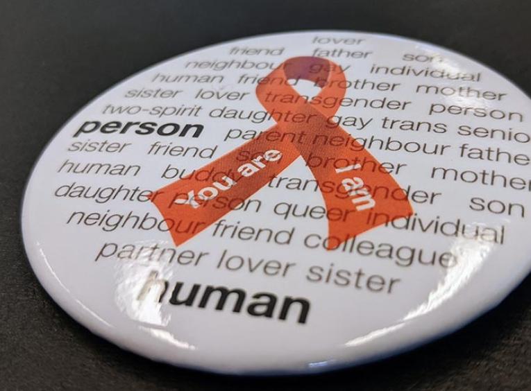 A World Aids Day button is pictured. The button features a red ribbon on a white background with words that humanize people battling HIV.