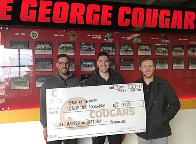 Three men holding a large cheque in front of a red sign Prince George Cougars.