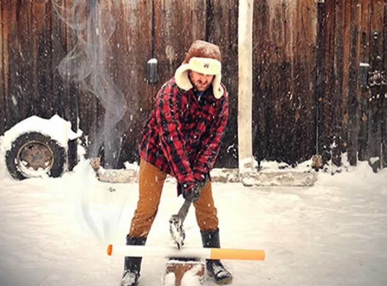Man wearing a plaid jacket and hat outside in the snow chopping a large pretend cigarette.