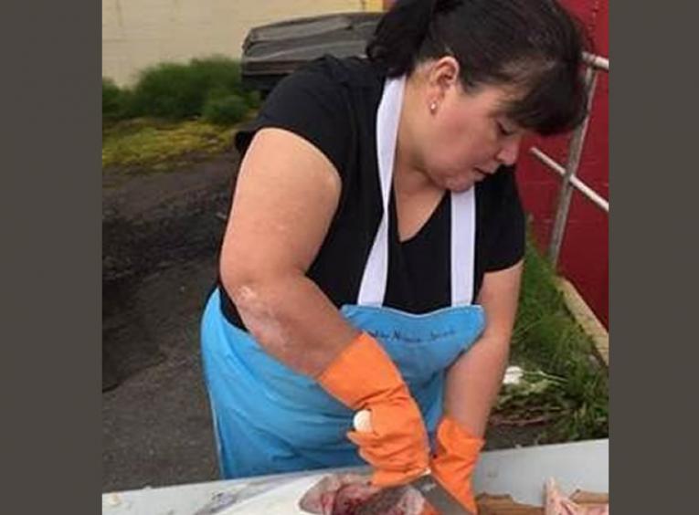Woman with dark hair and blue apron cleaning a fish.