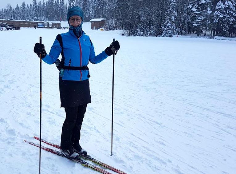 A woman on skis stands outside on the snow, holding ski polls and smiling into the camera. 
