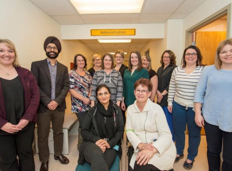 13 people stand in a hospital hallway, smiling at the camera. 