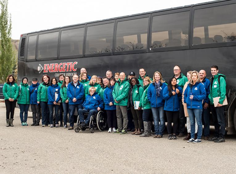A large group of people in blue or green BC Winter Games jackets stand in front of a bus.