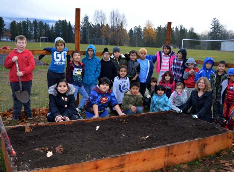 Elementary school children, some holding gardening tools, and a teacher stand in front of a planter box.