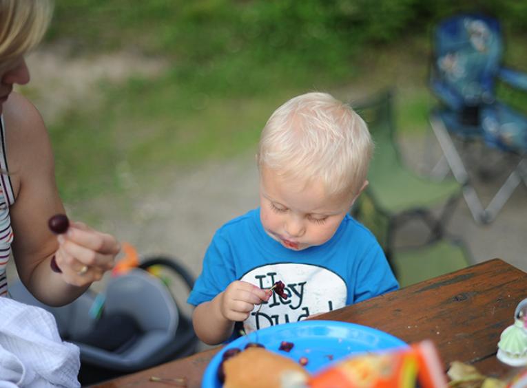 A toddler boy looks at a cherry he is eating at a picnic table while his mom looks at him. 