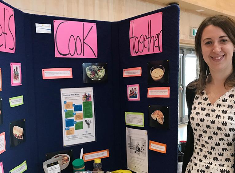 A smiling woman standing next to a large poster board titled "Let's Cook Together" with a variety of ingredients, including apples and peanut butter, sitting on the table in front of it.