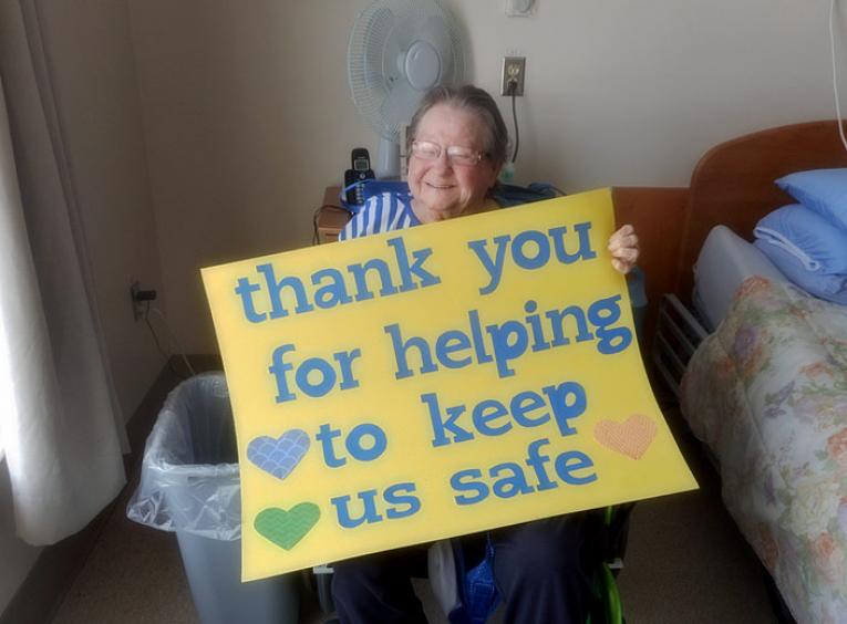 A female long-term care resident holds a sign that says, "thank you for helping to keep us safe." 