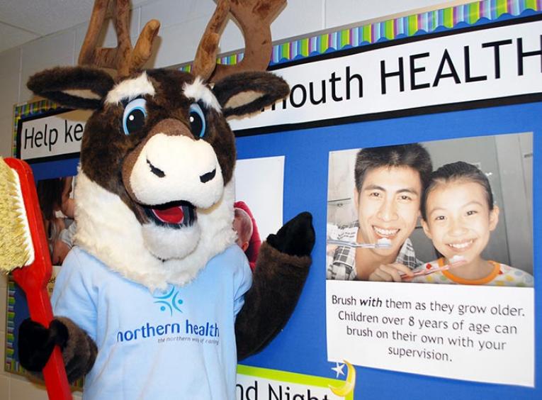Northern Health's caribou mascot holds a giant toothbrush in front of a display promoting mouth health. 