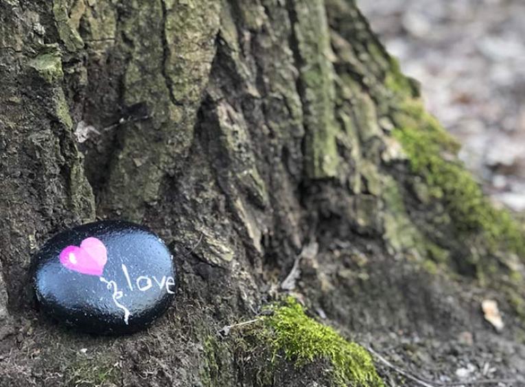 A small rock with a heart-shaped balloon and the word "love" painted on it lays against the base of a tree. 