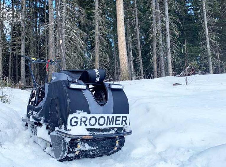 A trailer groomer sits on snow in a forest.