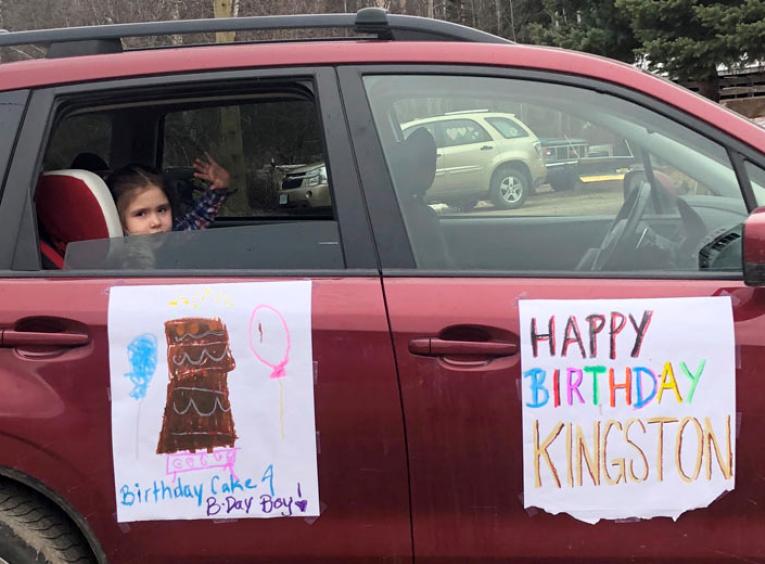 A young girl waves out the back of a red SUV decorated with homemade posters featuring birthday wishes. 