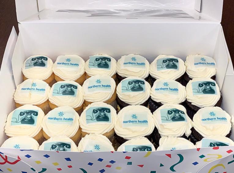 A box of cupcakes that are decorated with the Northern Health logo and pictures of old phones.