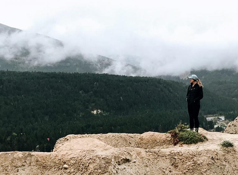 A woman stands at the edge of a cliff, forested mountains touching clouds in the background. 