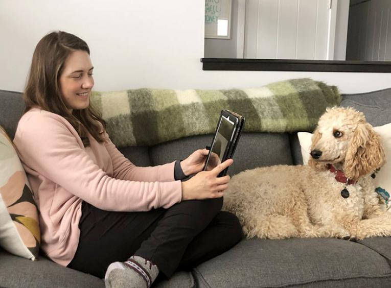 A young woman sits on a couch, chatting to another person on a tablet. A poodle sits on the other side of the couch.