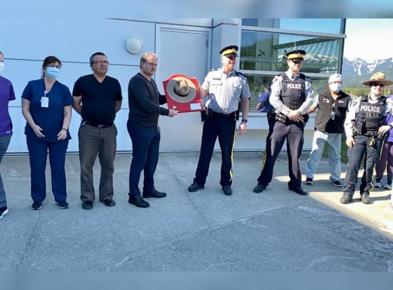 A group of health care staff and RCMP line up, with a man in the middle holding up a plaque with a hat on its front. 