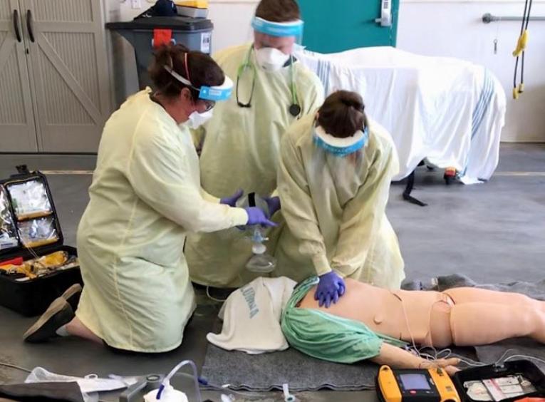 Three people wearing personal protective gowns and masks perform CPR on a simulation dummy. 