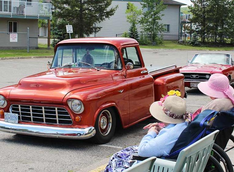 An old red pickup truck drives past two women in wheelchairs.