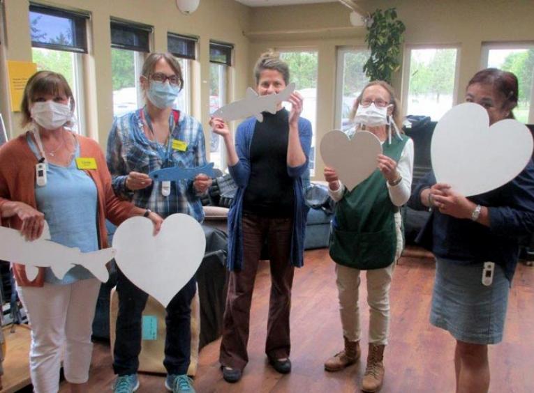Five women stand next to each other, several wearing masks, holding cut out hearts and fish.  