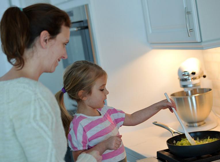 In a kitchen, a woman holds a young girl who stirs food in a pan on the stove.