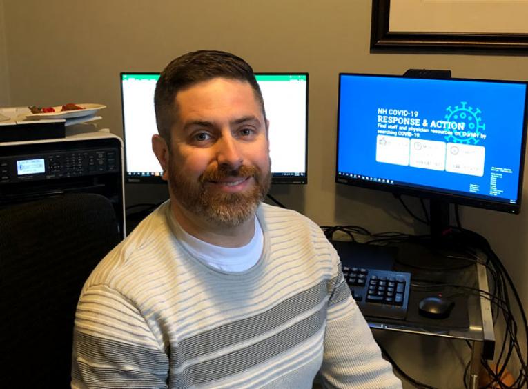 A man sits at his home office, two computer screens behind him.