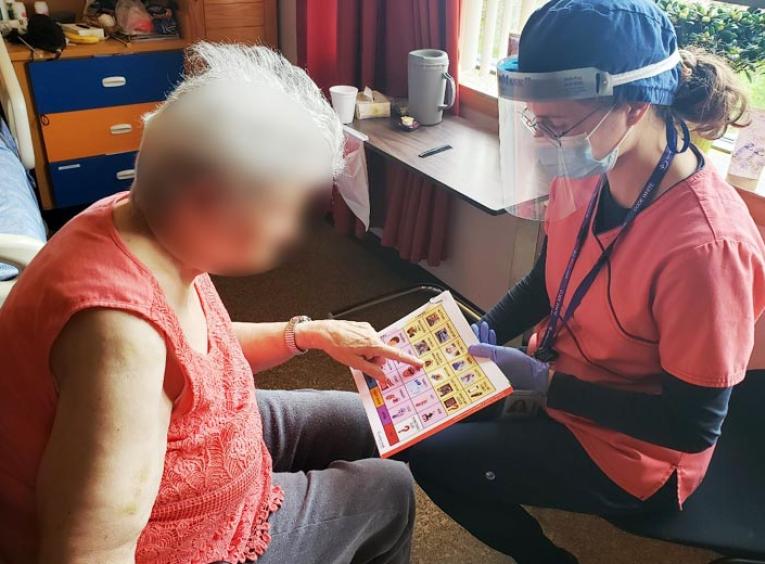 An elderly woman points to a booklet held by a person in personal protective equipment.