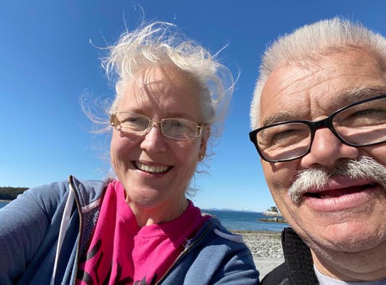 A man and woman on a rocky beach smile into the camera, blue sky and water behind them.