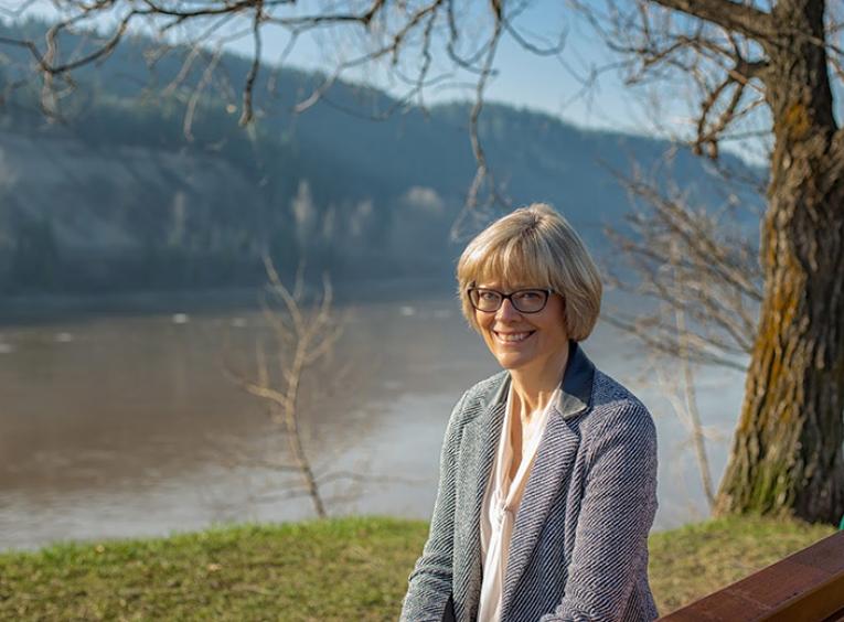 A smiling woman sits on a bench above a river.