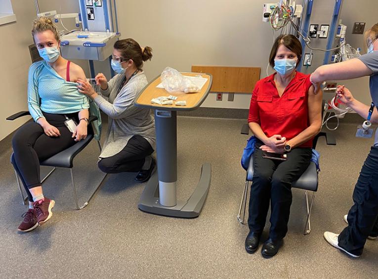 Two women are sitting in a hospital setting receiving vaccinations from nurses.